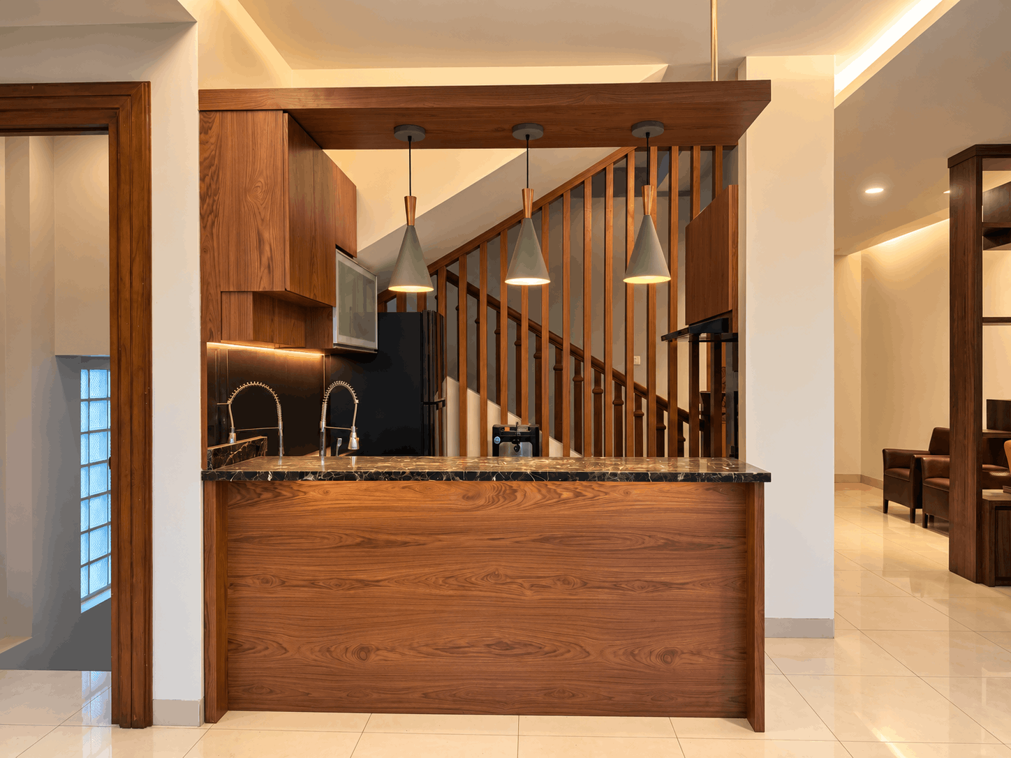 Pantry modern dengan kitchen bar finishing kayu walnut, countertop marmer gelap, lampu gantung minimalis, dan tangga kayu di belakang.