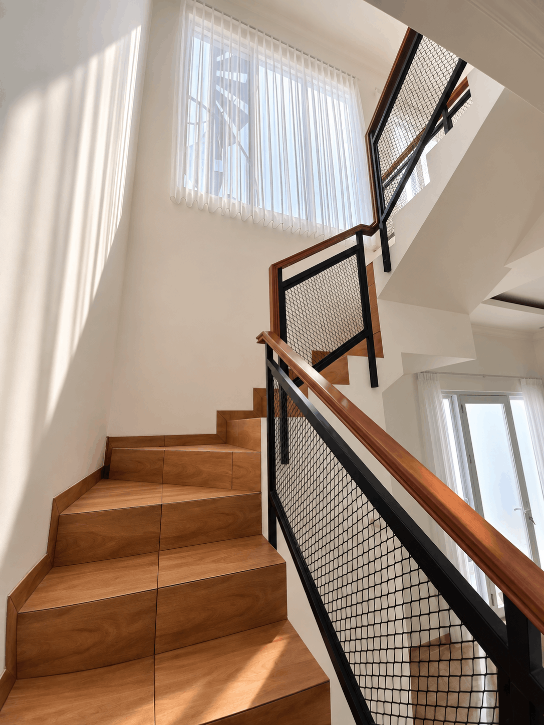 Tangga rumah modern dengan finishing motif kayu, railing besi hitam bermotif mesh, handrail kayu, dan pencahayaan alami dari jendela besar.