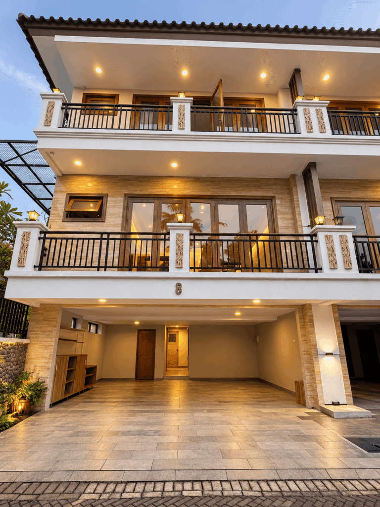 Fasad rumah mewah bertingkat dengan balkon luas, railing hitam, pencahayaan eksterior hangat, dan area carport lebar.
