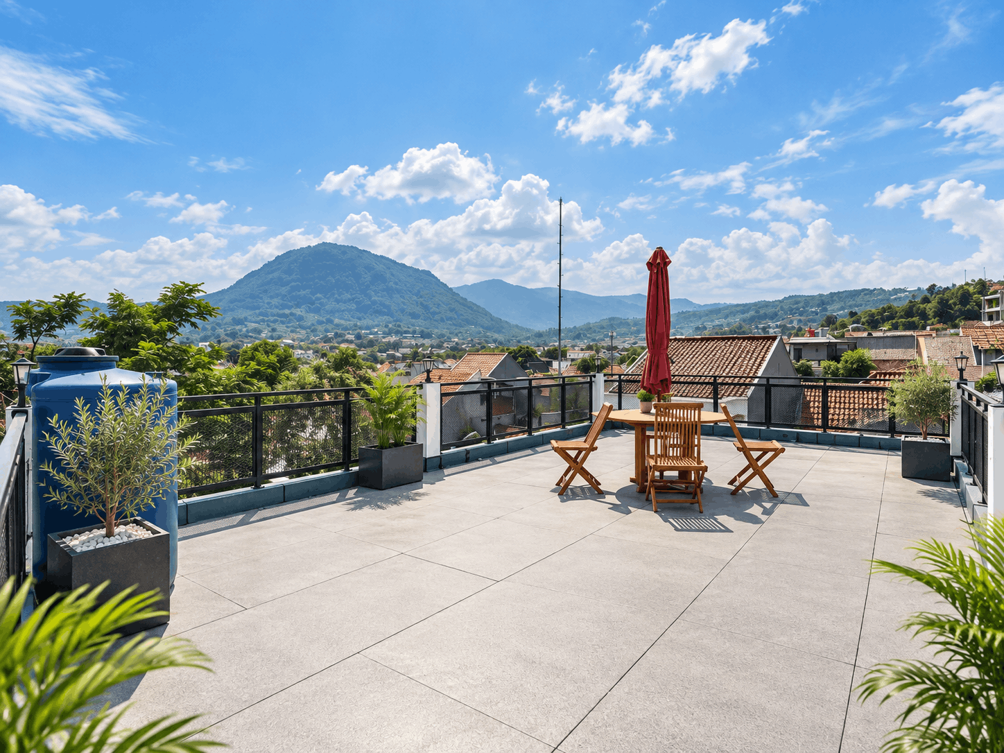 Area rooftop rumah dengan meja kursi outdoor, railing hitam, tanaman dekoratif, dan pemandangan pegunungan